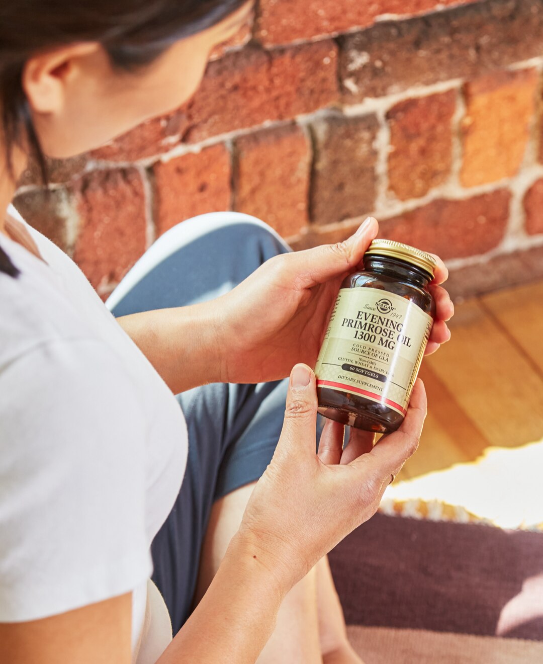 Overlooking right shoulder of woman holding Solgar evening primrose oil supplement in a sunny room beside an exposed brick wall