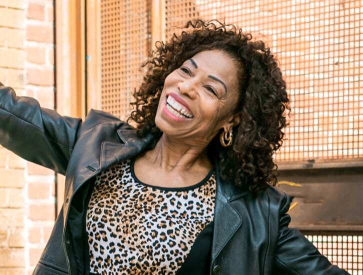 A Black, middle-aged woman smiling, wearing a leopard-print shirt, black leather jacket, with her arms outstretched and a black hat in one hand.