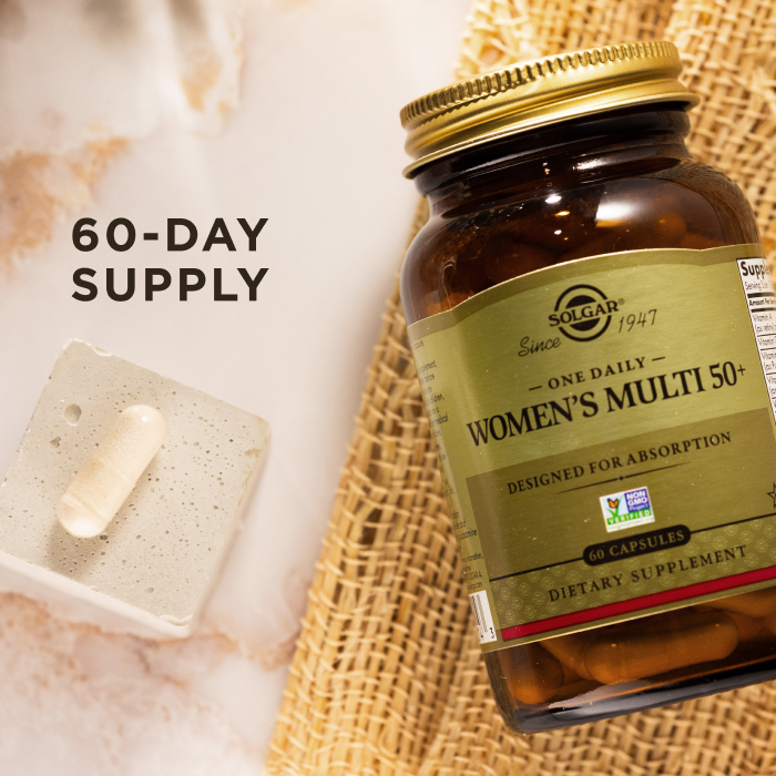 A bird's eye view of an amber glass bottle of Solgar One Daily Women's Multi 50+ supplement on a marble countertop. Beside the glass bottle is a single capsule showcasing the size and scale of the supplement. The image says, "60-day supply".