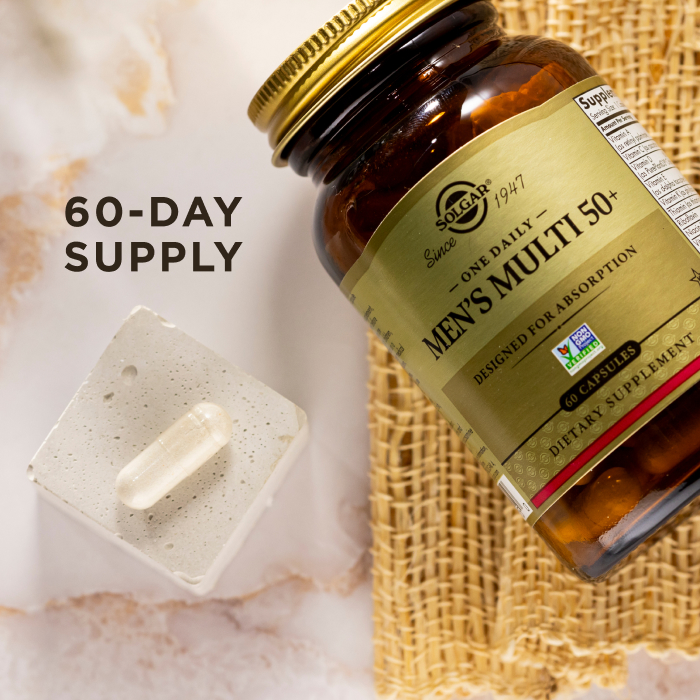 A bird's eye view of an amber glass bottle of Solgar One Daily Men's Multi 50+ supplement on a marble countertop. Beside the glass bottle is a single capsule showcasing the size and scale of the supplement. The image says, "60-day supply".