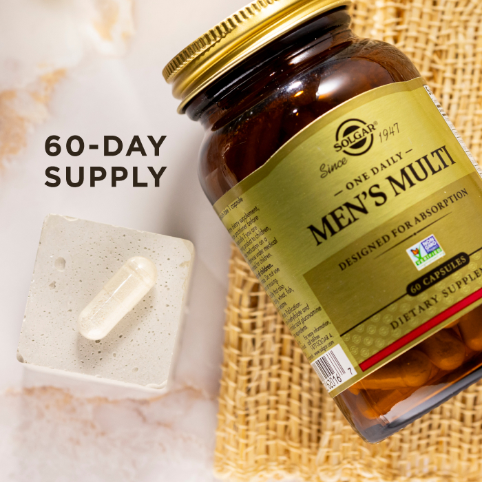 A bird's eye view of an amber glass bottle of Solgar One Daily Men's Multi supplement on a marble countertop. Beside the glass bottle is a single capsule showcasing the size and scale of the supplement. The image says, "60-day supply".