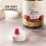 A single serving of Solgar Ultra Potency Zinc Gummies sitting on a white stone riser on a marble countertop. The gummy and riser is in focus, the bottle of vitamins is visible but blurred in the background. The image says, "60-day supply".