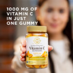 A woman holding a bottle of Solgar Ultra Potency Vitamin C Gummies towards the camera. The bottle is in focus but the woman is blurred with the camera's depth of field. The image says, "1000 mg of Vitamin C in just one gummy."