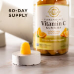 A single serving of Solgar Ultra Potency Vitamin C Gummies sitting on a white stone riser on a marble countertop. The gummy and riser is in focus, the bottle of vitamins is visible but blurred in the background. The image says, "60-day supply".