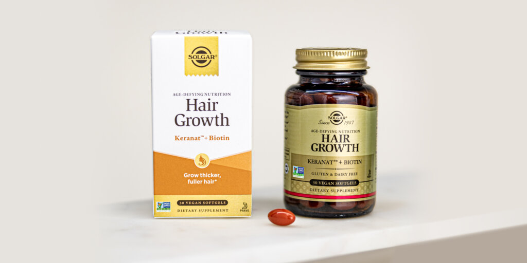 A box & bottle of Solgar® Hair Growth on a white marble shelf. One capsule from the bottle is placed in front to showcase the shape, size, and color. 