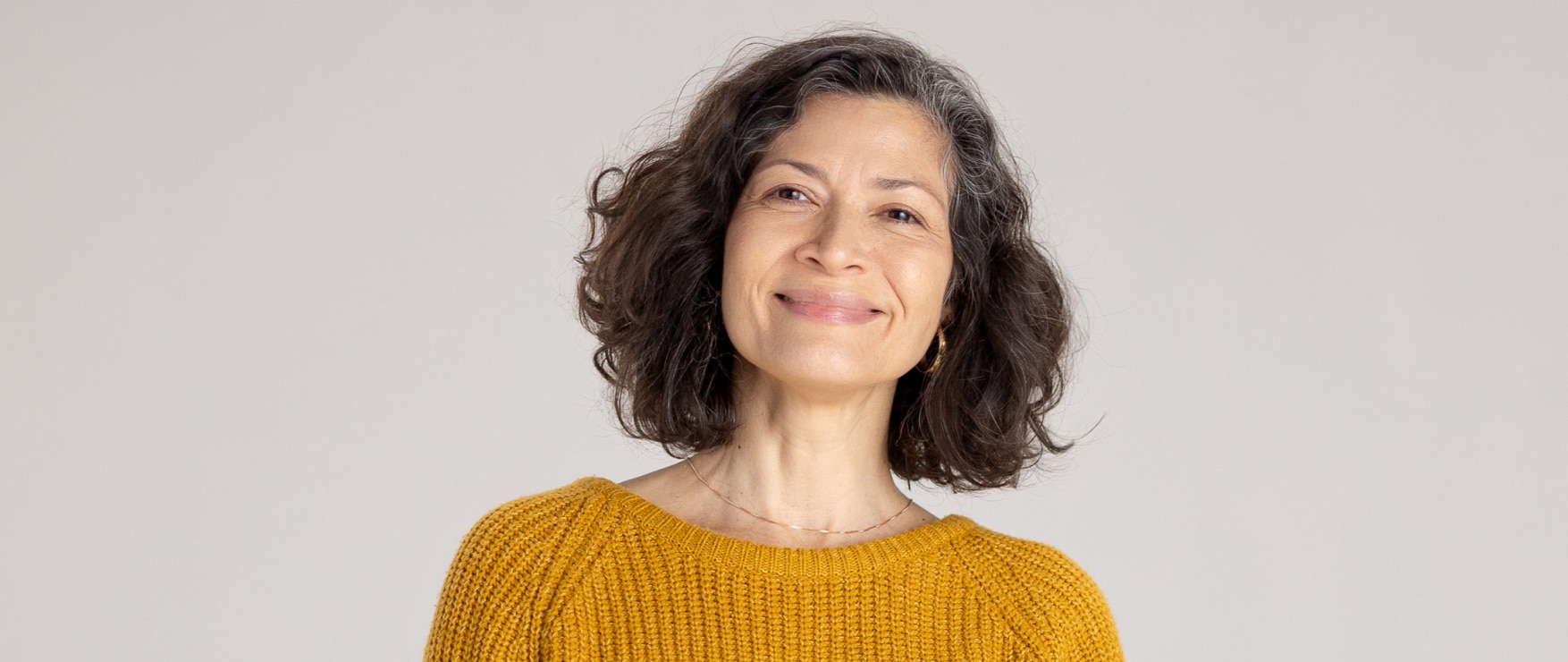 A smiling and confident middle-aged woman with thick, curly hair and glowing skin wearing a gold colored sweater, standing in front of a warm white background.
