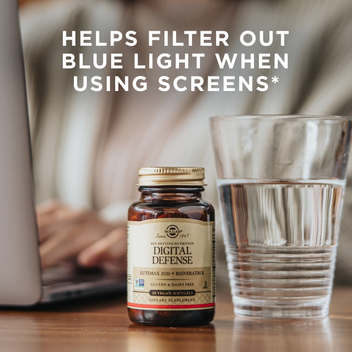 A woman working at a laptop with a glass of water and a bottle of Solgar Digital Defense beside her. Text on image reads, "Helps filter out blue light when using screens*"