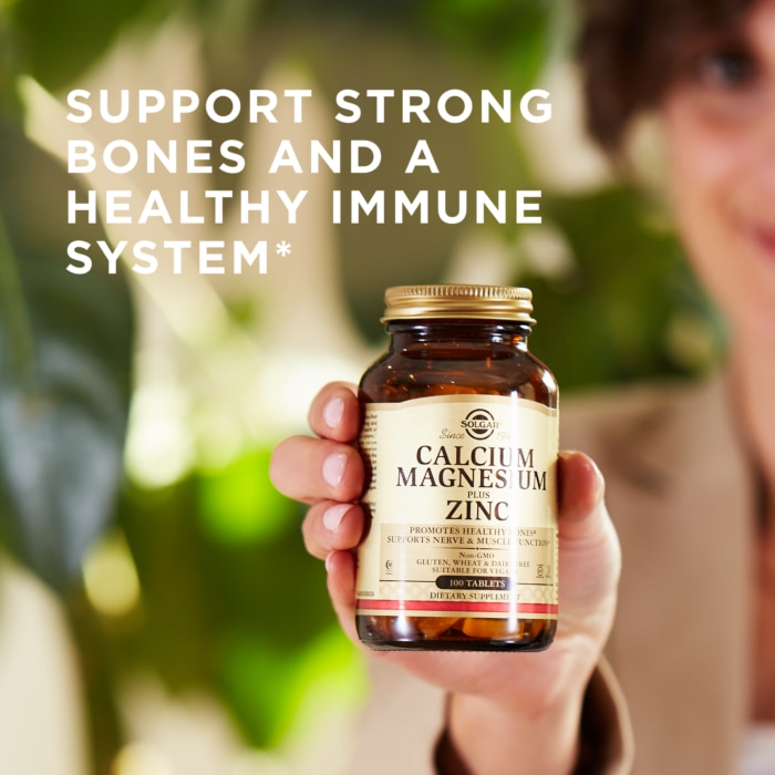 A woman holds a bottle of Solgar's Calcium Magnesium Plus Zinc Tablets in the foreground. Text reads "support strong bones and a healthy immune system"