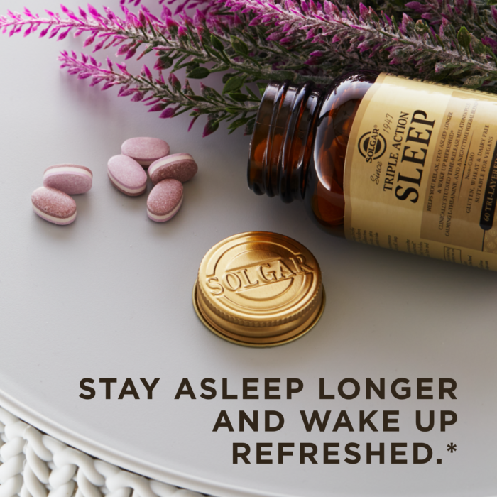 An amber glass bottle of Solgar's Triple Action Sleep Tri-layer tablets laying on its side with it's golden lid removed and placed next to it. A pile of 6 of the tablets lays next to a sprig of lavender on a flat white surface. Text overlaid reads "Stay asleep longer and wake up refreshed.*"