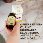 An outstretched hand holds an amber glass bottle of Solgar's Ester-C® Plus Immune Complex softgels above its outer packaging on a marble kitchen surface. A text overlay below reads: "Offers Ester-C®, zinc, echinacea, elderberry, astagralus, and more."