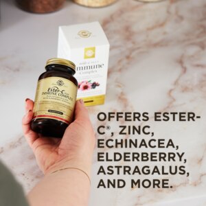 An outstretched hand holds an amber glass bottle of Solgar's Ester-C® Plus Immune Complex softgels above its outer packaging on a marble kitchen surface. A text overlay below reads: "Offers Ester-C®, zinc, echinacea, elderberry, astagralus, and more."