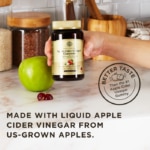 A hand holds an amber glass bottle of Solgar Apple Cider Vinegar Gummies above a kitchen surface next to an apple and some of the loose gummies. A text overlay reads "Made with liquid apple cider vinegar from US-grown apples." Another stamp on the image reads "Better taste than the #1 Apple Cider Vinegar Gummy."