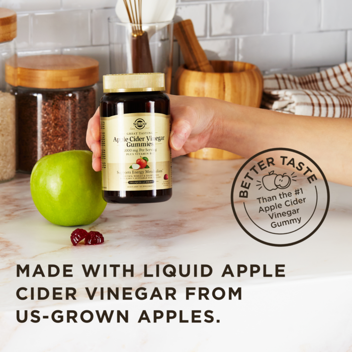 A hand holds an amber glass bottle of Solgar Apple Cider Vinegar Gummies above a kitchen surface next to an apple and some of the loose gummies. A text overlay reads "Made with liquid apple cider vinegar from US-grown apples." Another stamp on the image reads "Better taste than the #1 Apple Cider Vinegar Gummy."