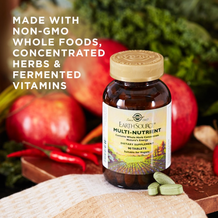 A bottle of Solgar's Earth Source® Multi-Nutrient Tablets sits next to apples and chili peppers. Text reads "made with non-GMO whole foods, concentrated herbs, and fermented vitamins"