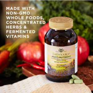 A bottle of Solgar's Earth Source® Multi-Nutrient Tablets sits next to apples and chili peppers. Text reads "made with non-GMO whole foods, concentrated herbs, and fermented vitamins"