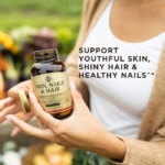 A bottle of Solgar's Skin, Nails & Hair Tablets held in front of a woman in a white top. Text reads "support youthful skin, shiny hair and healthy nails*"