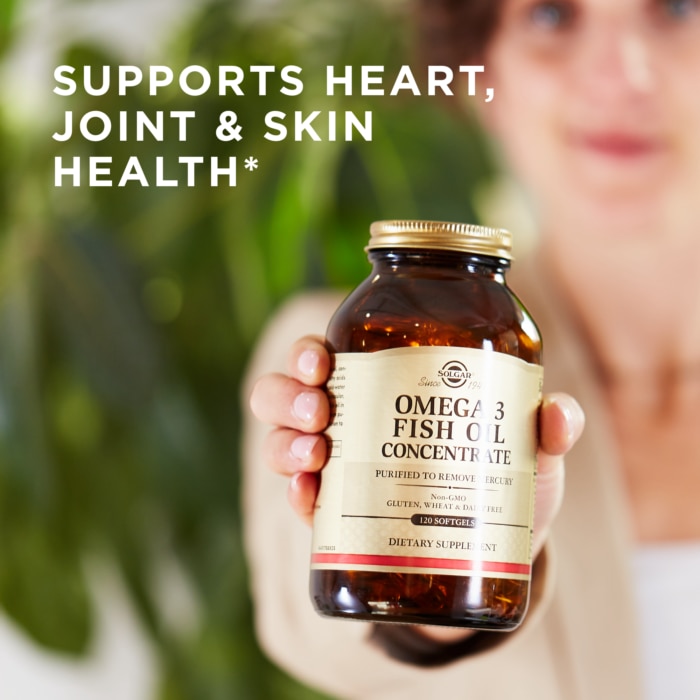 A woman holds up Solgar's Omega-3 Fish Oil Concentrate Softgels in an amber glass bottle. Text reads 'supports heart, joint, and skin health"