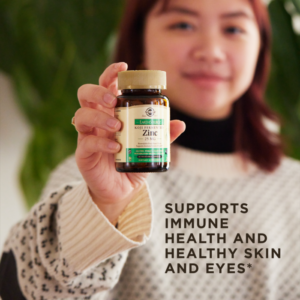 A smiling woman holds a bottle of Solgar's Earth Source Koji Fermented Zinc Vegetable Capsules up to the camera. Text overlaid reads 'supports immune health and healthy skin and eyes'