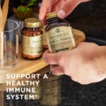 Solgar's Vitamin D3 Cholecalciferol softgels held in a persons hands, with other Solgar products on a table in the background. Text reads "Support a healthy immune system*"