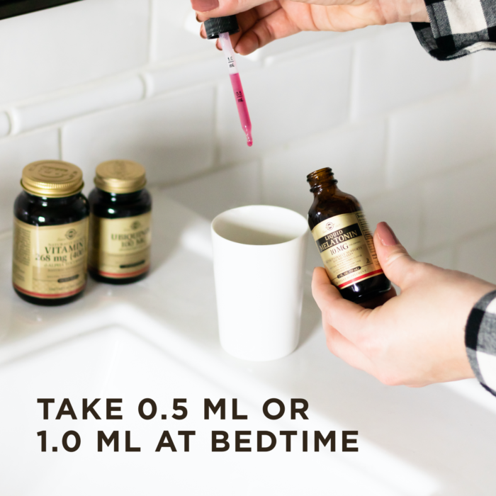 A person uses the dropper lid of Solgar's Liquid Melatonin 10 mg - Natural Black Cherry Flavor to drop the liquid into a white cup. Other Solgar products are in the background on a white surface. Text reads "take 0.5ml or 1ml at bedtime.